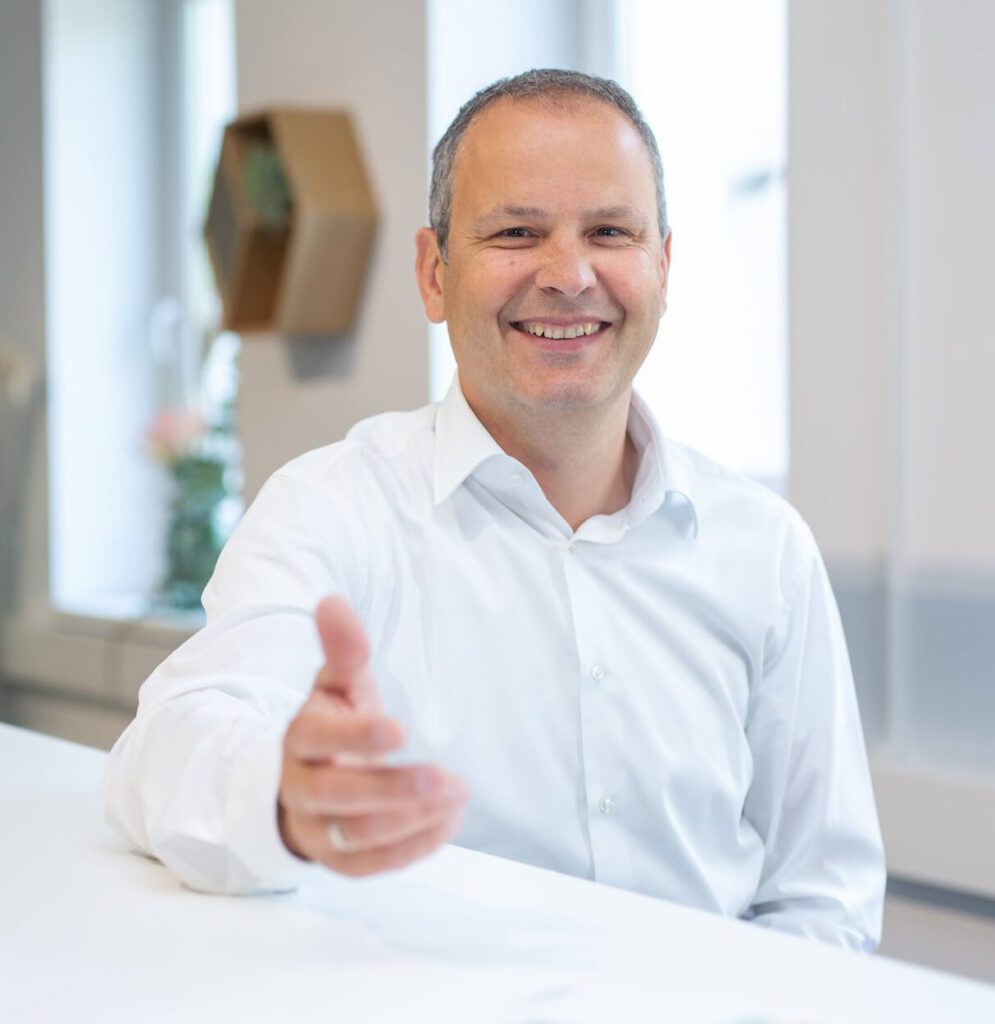 Ein Mann in einem weißen Hemd lehnt lächelnd an einem weißen Tisch